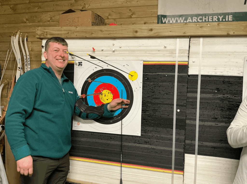 A man with a green shirt pointing to a center-hit on an indoor archery target