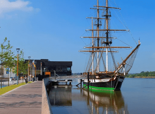 Large three-masted wooden tall ship with white sails docked at a river quay